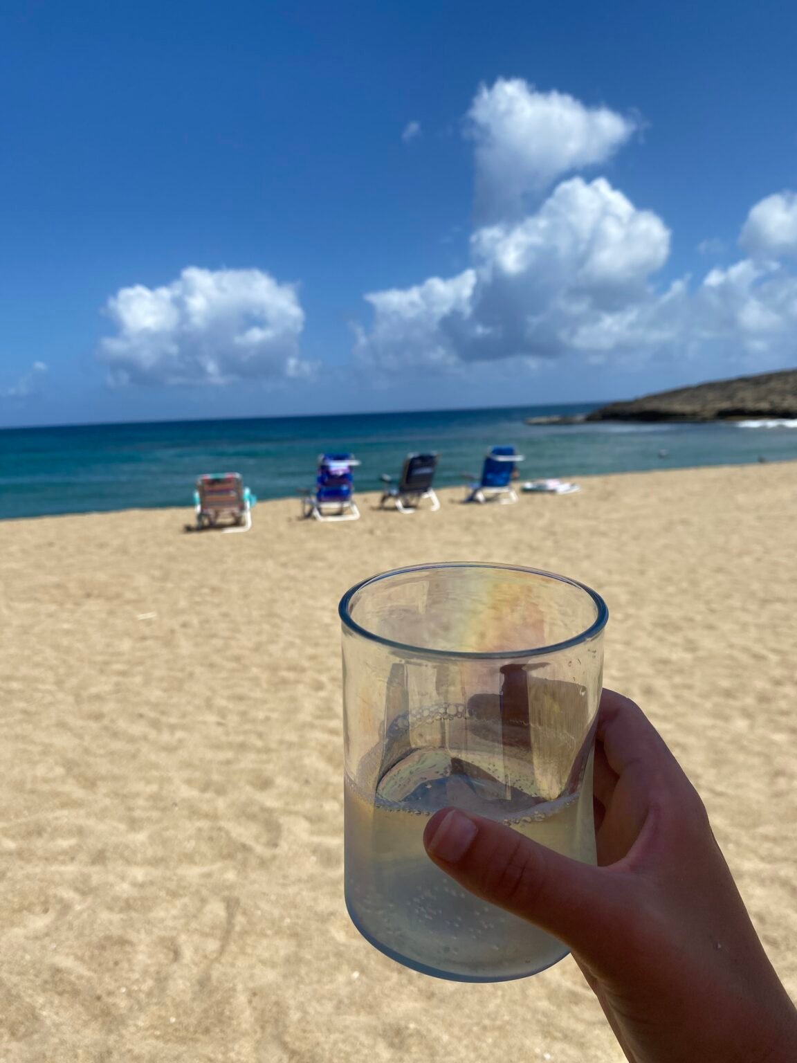 una mano sosteniendo un vaso con vino dentro y en el fondo cuatro sillas de playas alineadas y la playa montones 