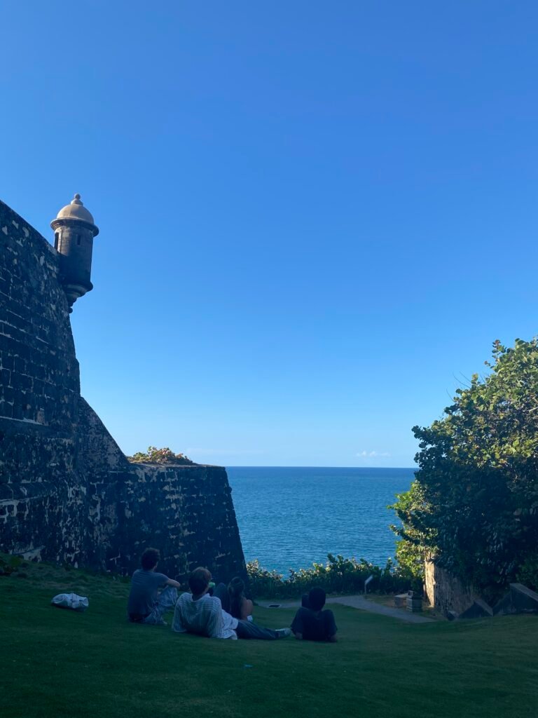 cuatro jovenes están sentados en la grama del Morro con vista hacia el Océano Atlántico