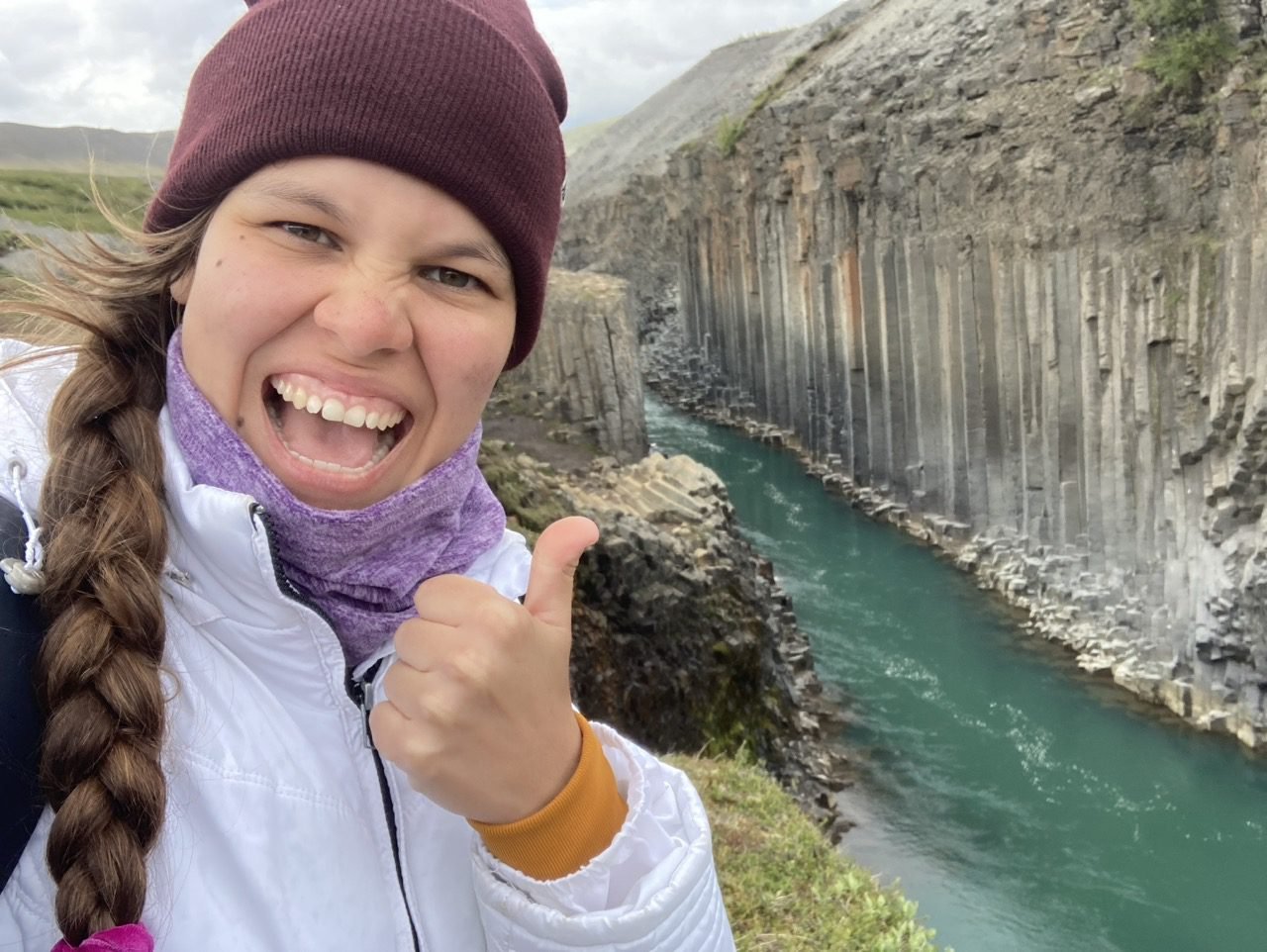 La joven sonríe a la cámara y en el fondo se puede apreciar el cañón Studlagil de Islandia