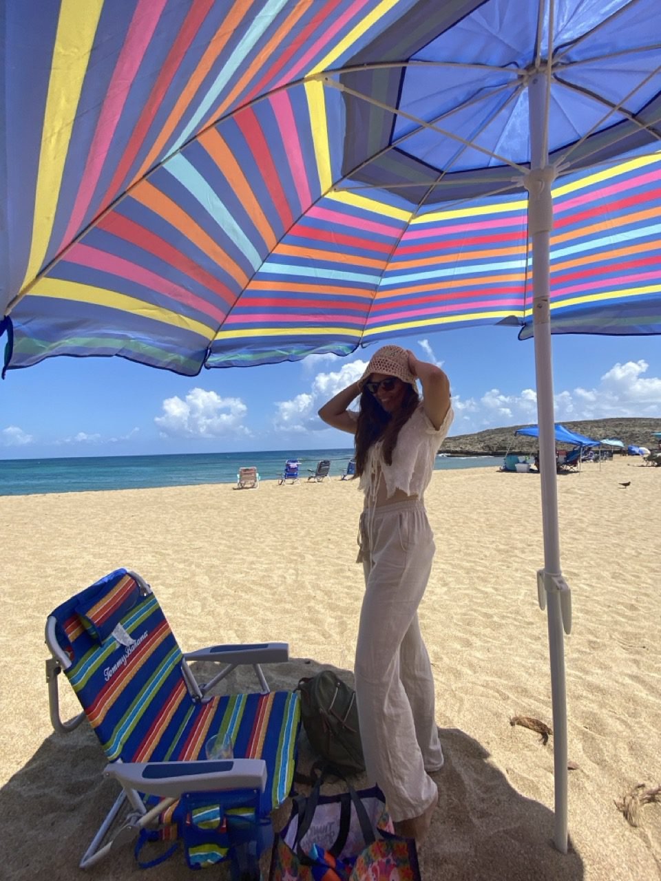 mujer de pie bajo una sombrilla de playa de colores y aguantando su gorro de playa para que el viento no se lo lleve